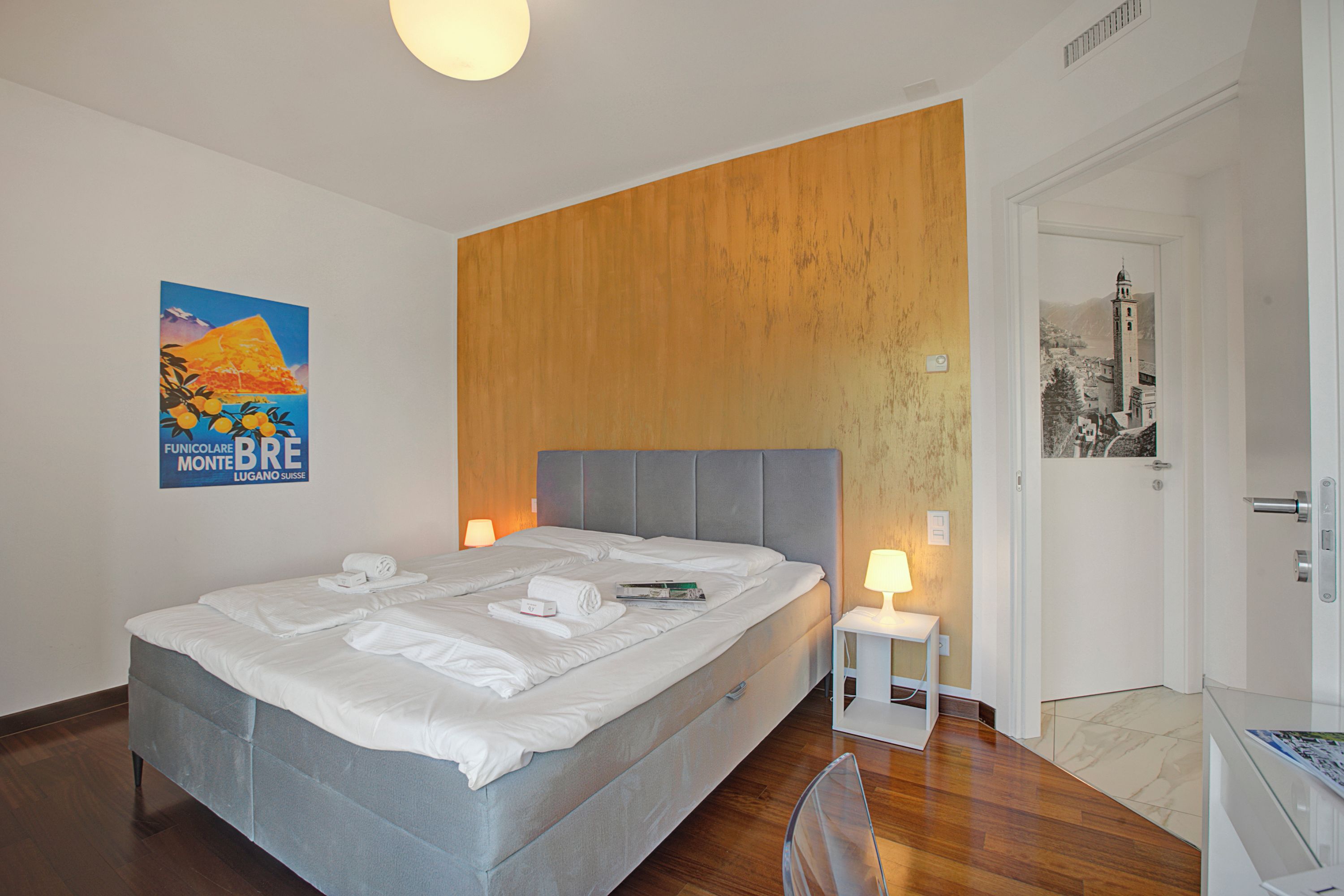 Uma cama bem arrumada com lençóis brancos, uma pequena mesa de cabeceira com abajur e um cartaz na parede.