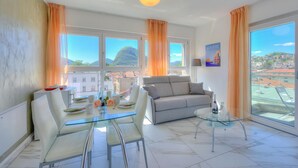 A modern living room with a glass table, white chairs, a grey sofa, and large windows with curtains. - Attico al Parco: Penthouse in Lugano Balcony, A/C & 2 min From Lake (Lugano)