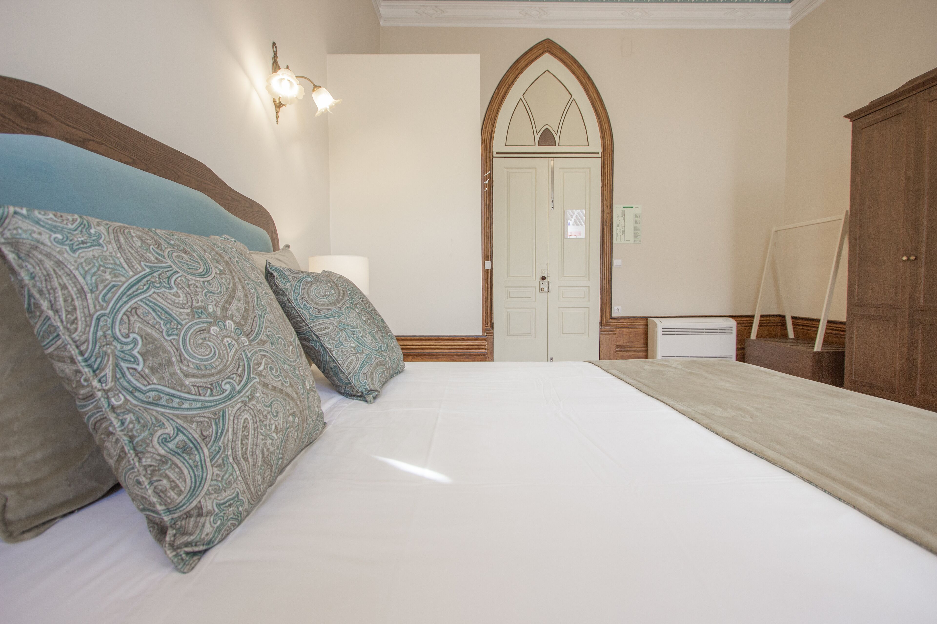 Un dormitorio con una cama grande, almohadas estampadas, un cabecero de madera, una lámpara de pared y un armario de madera con un arco.