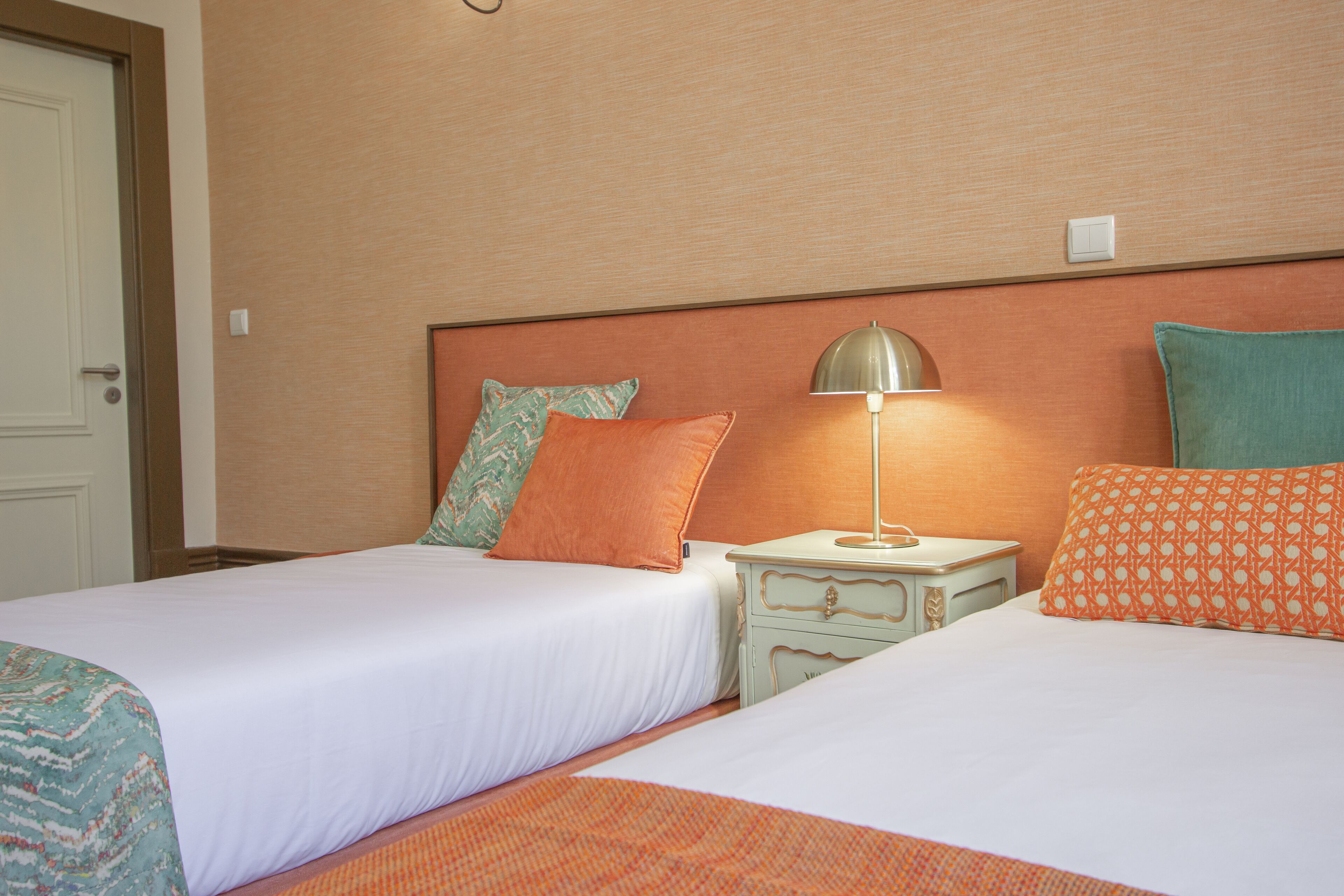Habitación de hotel con dos camas, cada una con cubrecama blanco y varias almohadas. Hay una mesita de noche con lámpara entre las camas. La habitación tiene cabecera de madera y una puerta a la izquierda.