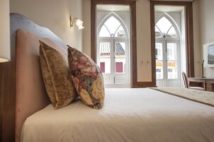 Un dormitorio con una cama grande, dos almohadas estampadas con motivos florales, un cabecero de madera y vistas a un edificio desde la ventana.