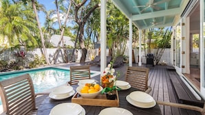 Terraza de madera con piscina, sillas de descanso y zona de comedor con platos y fruta.