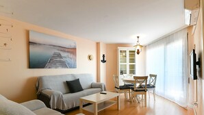 Ein Wohnzimmer mit einer Couch, einem Couchtisch und einem Essbereich mit Stühlen und einem Tisch. An der Wand hängt ein Gemälde, eine Chandelieber ist angebracht und ein Fernseher ist wandmontiert.