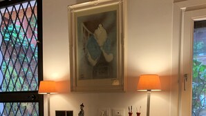 A room with a wooden desk, two lamps, a framed painting, and a window with a view of greenery. - Small Luxury Apartment (Venezia)