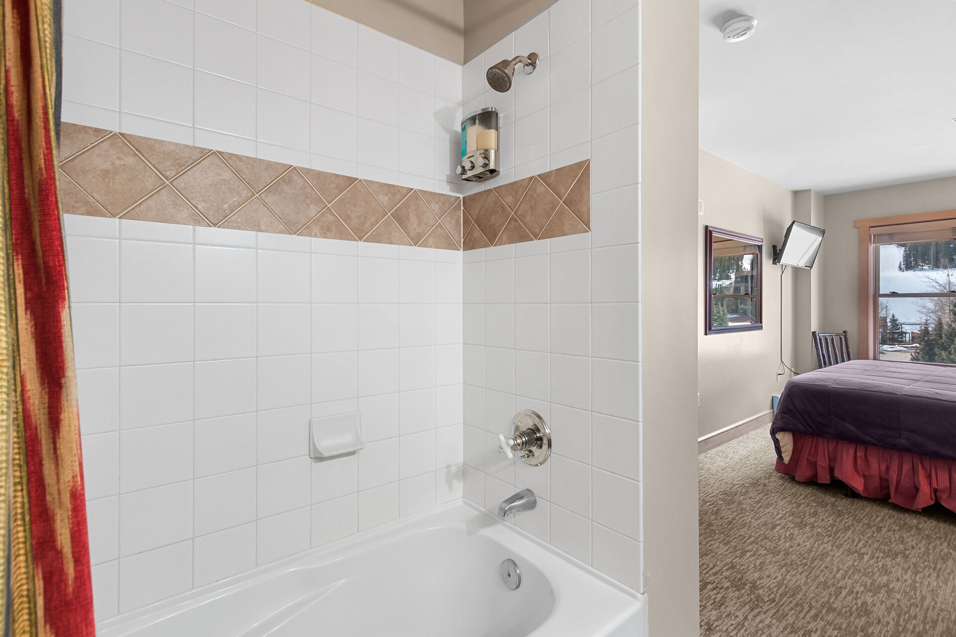A bathroom with white tiles, a bathtub, a showerhead, and a window with a view of trees.