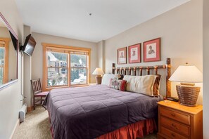 A bedroom with a large bed, wooden headboard, two bedside lamps, a chair, a TV mounted on the wall, and a window with a view of buildings and trees. - 1 Minute Walk to the Super Bee Chair! Enjoy the Best of Copper from this Beautif (Frisco)