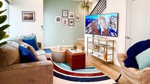 A living room with a sofa, ottoman, and a coffee table. There is a TV mounted on the wall and a staircase in the background.
