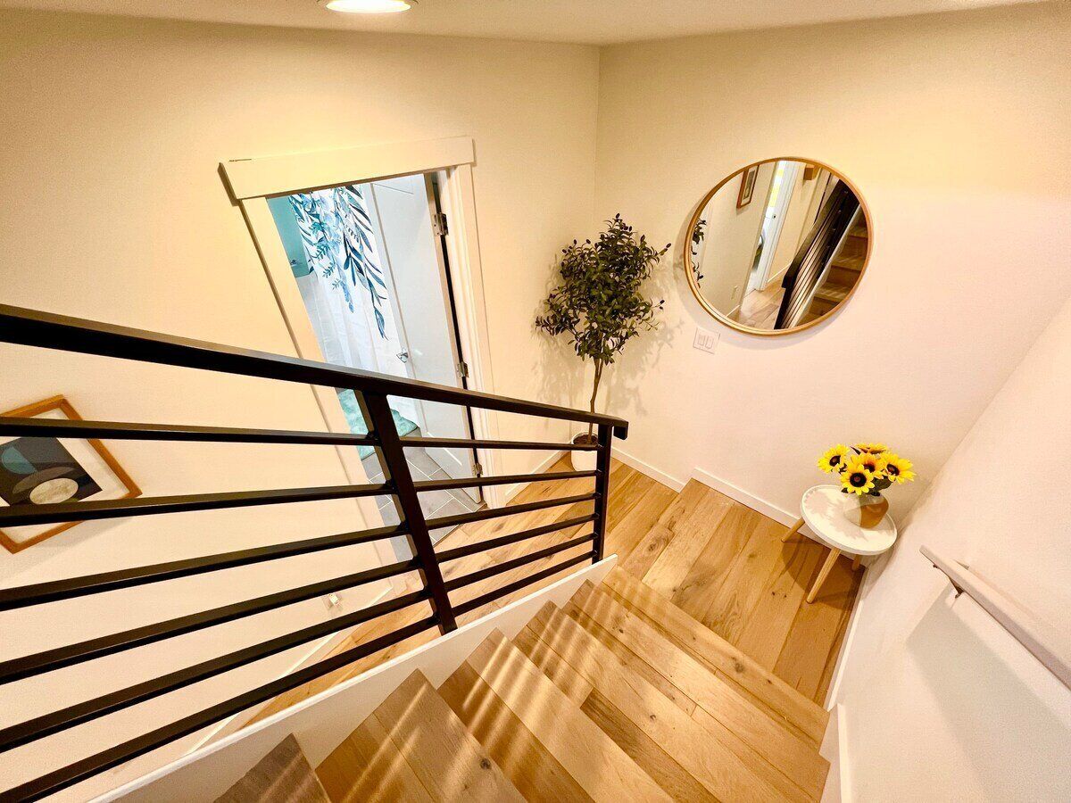 Un escalier avec une rampe noire, un plancher en bois, un miroir au mur et une petite table sur laquelle est posé un vase rempli de tournesols.