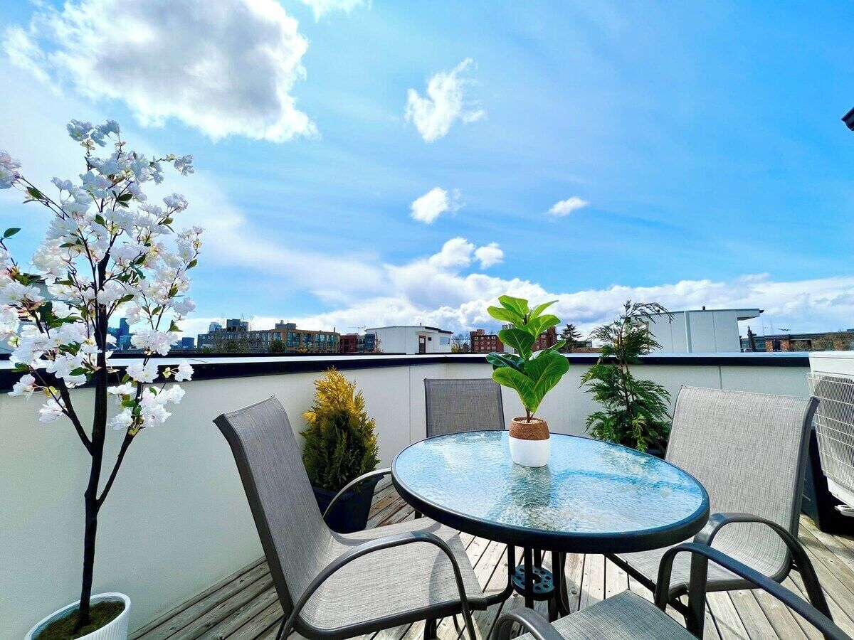 Une table ronde en verre, entourée de quatre chaises, se trouve sur une terrasse en toit, agrémentée de plantes en pot et offrant une vue sur le paysage urbain.