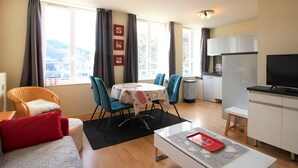 A modern, well-lit living room with a sofa, a dining table with chairs, a kitchen area with a refrigerator and a TV, and large windows with curtains. - La Bourboule: T2 bis Apartment for 5 ppl, 49m², with Wifi (La Bourboule)