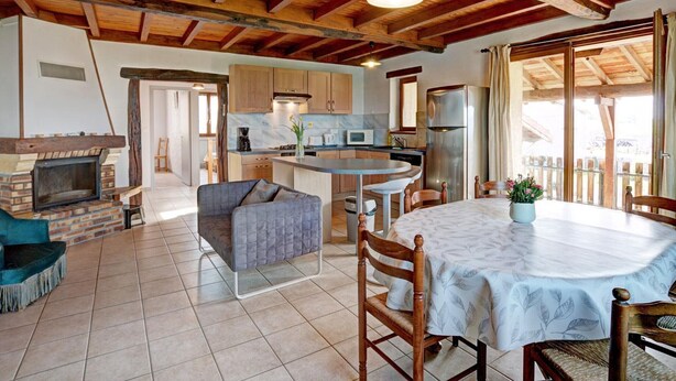 Een rustiek interieur met zichtbare houten balken, een open haard, een eetgedeelte met een ronde tafel, en een keuken met moderne apparatuur.