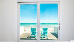 Blick durch eine Glastür auf einen Strand mit zwei türkisfarbenen Stühlen auf einem Holzdeck.
