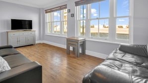 A living room with a sofa, a TV, a small table, and large windows with a view of the beach.