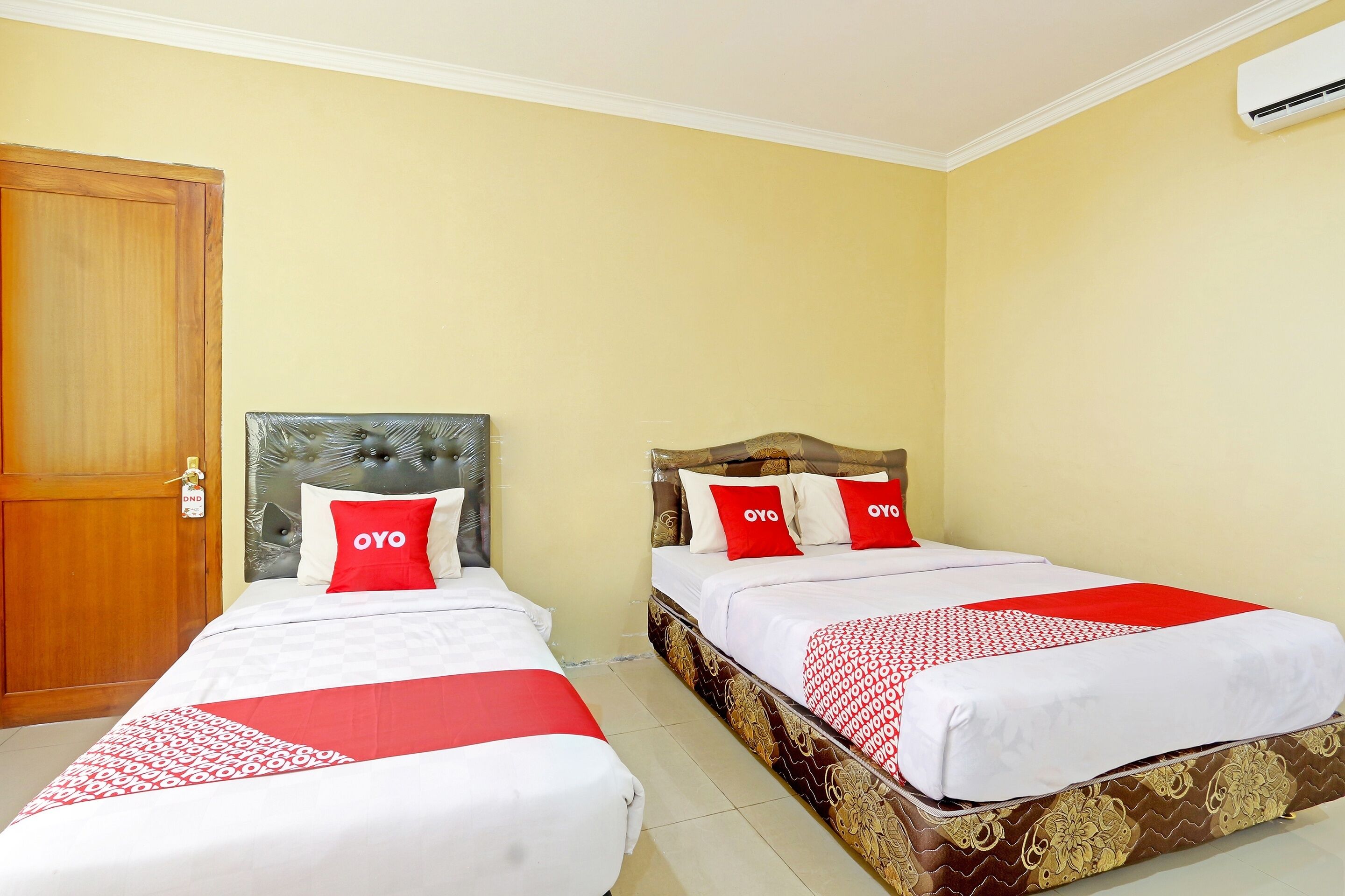 Une chambre d’hôtel avec deux lits, chacun recouvert d’une courtepointe rouge et blanche et agrémenté d’oreillers, une porte en bois et un climatiseur fixé au mur.