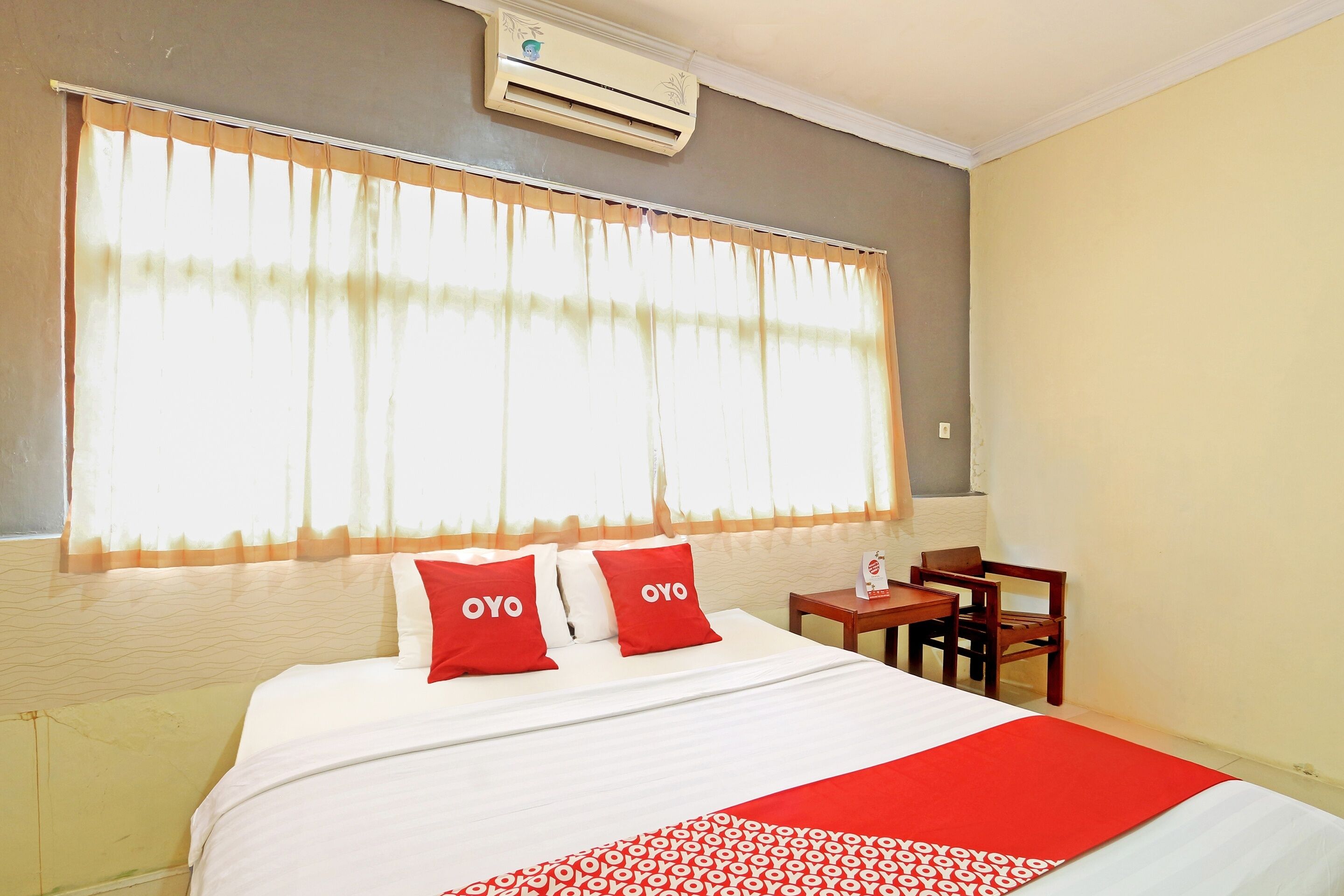 A hotel room with a bed, two pillows with "OYO" branding, a bedside table with a bottle, and a window with sheer curtains.