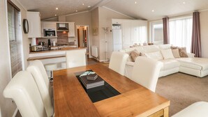 A spacious living area with a wooden dining table, white leather sofa, and a kitchen in the background.