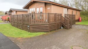 A wooden cabin with a fenced deck, a staircase, and a gravel driveway.