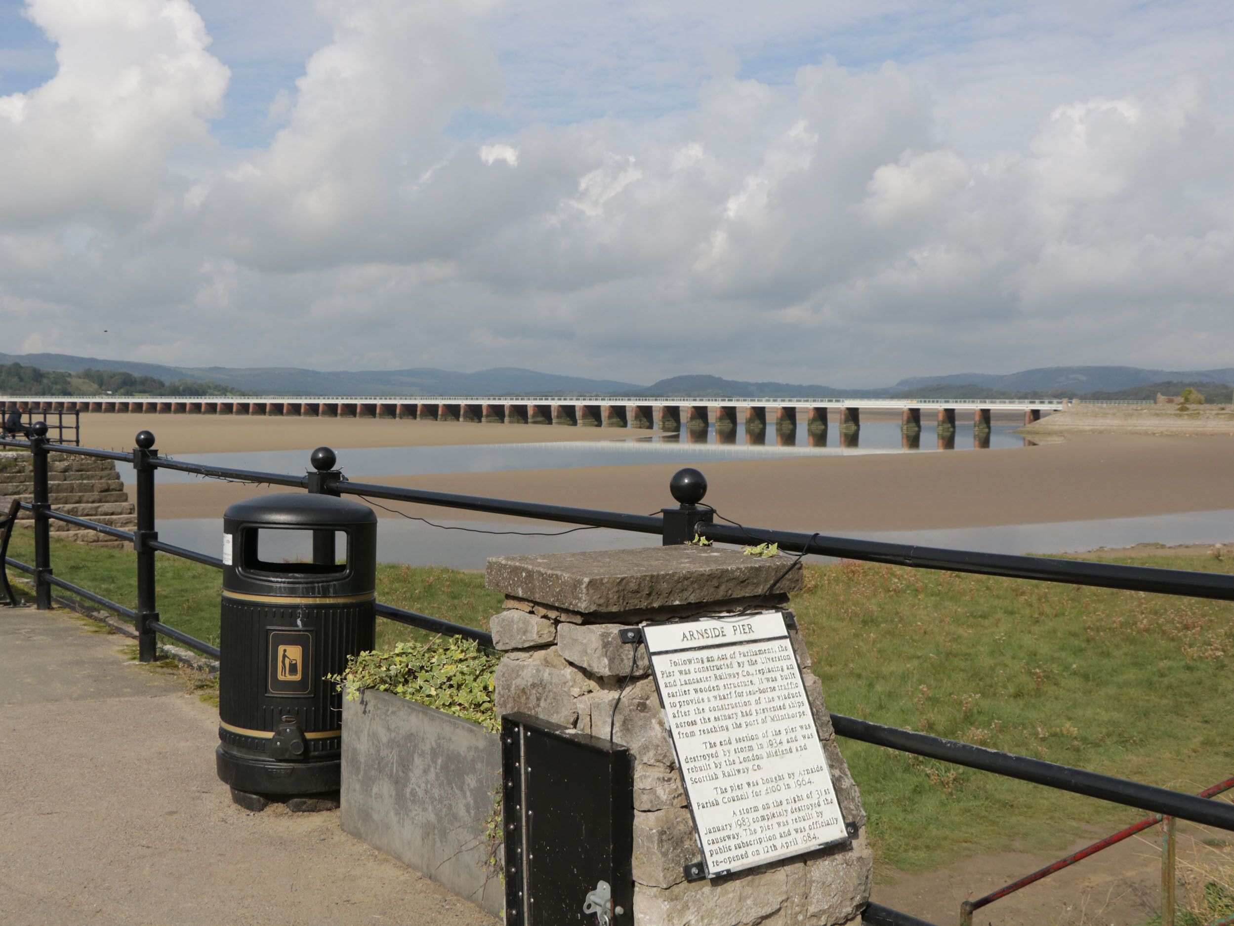 A pier with a historical plaque, a trash bin, and a view of a bridge over a river.