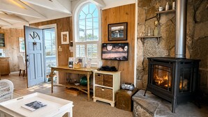 A cozy living room with a fireplace, wooden walls, a large window, and a television. - Fisherman's Cottage~Bodega Bay~Walk to Beach~Surf~Sand~Summer Coastal Escape! (Bodega Bay)