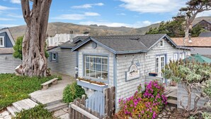 A small, single-story cottage with a large window and a porch, surrounded by a garden with pink flowers and a tree. - Fisherman's Cottage~Bodega Bay~Walk to Beach~Surf~Sand~Summer Coastal Escape! (Bodega Bay)