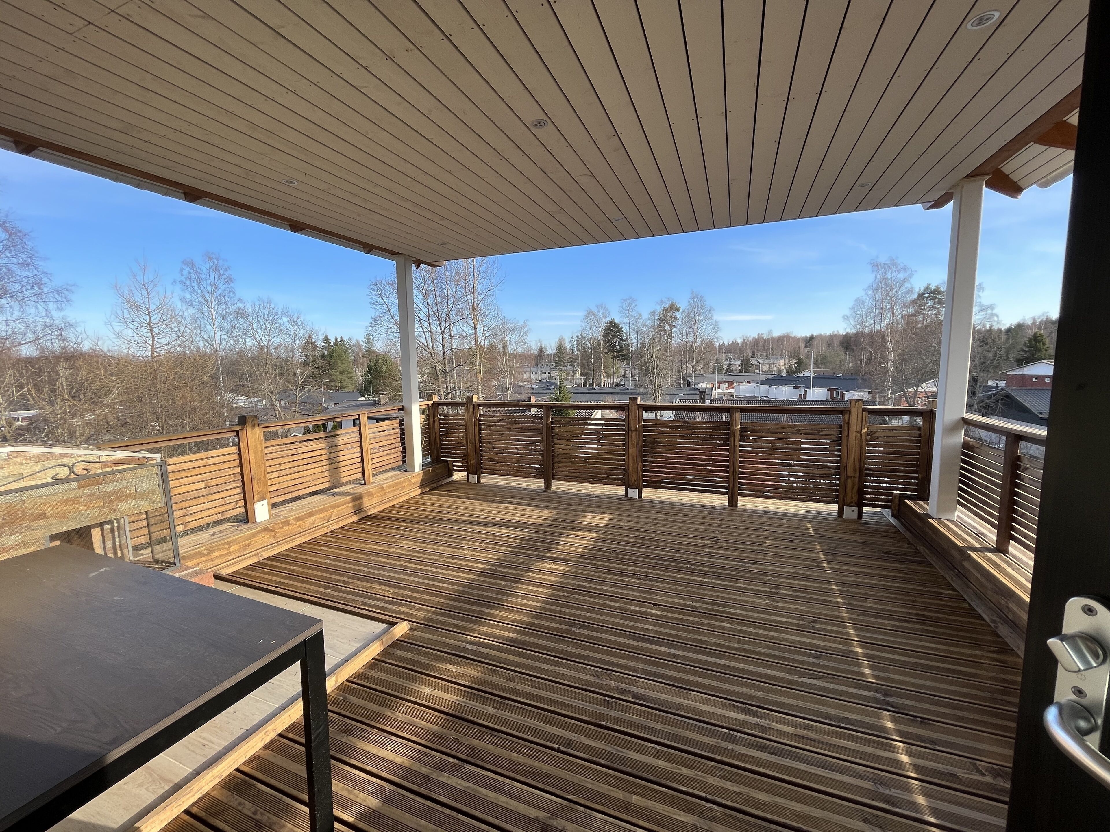 Une terrasse extérieure couverte, dotée d’un plancher en bois, d’une rambarde en métal et offrant une vue sur un quartier résidentiel.
