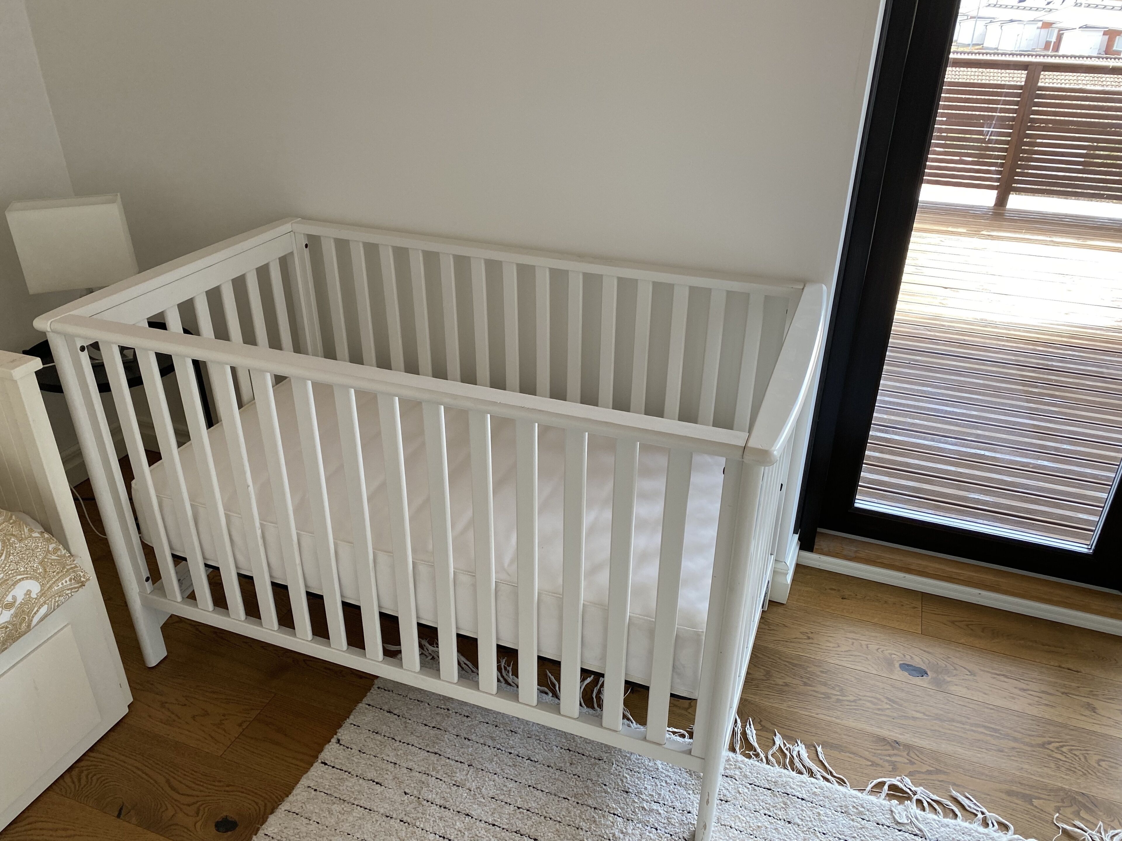 A white crib in a room with a wooden floor, a white cabinet, and a view through a glass door.