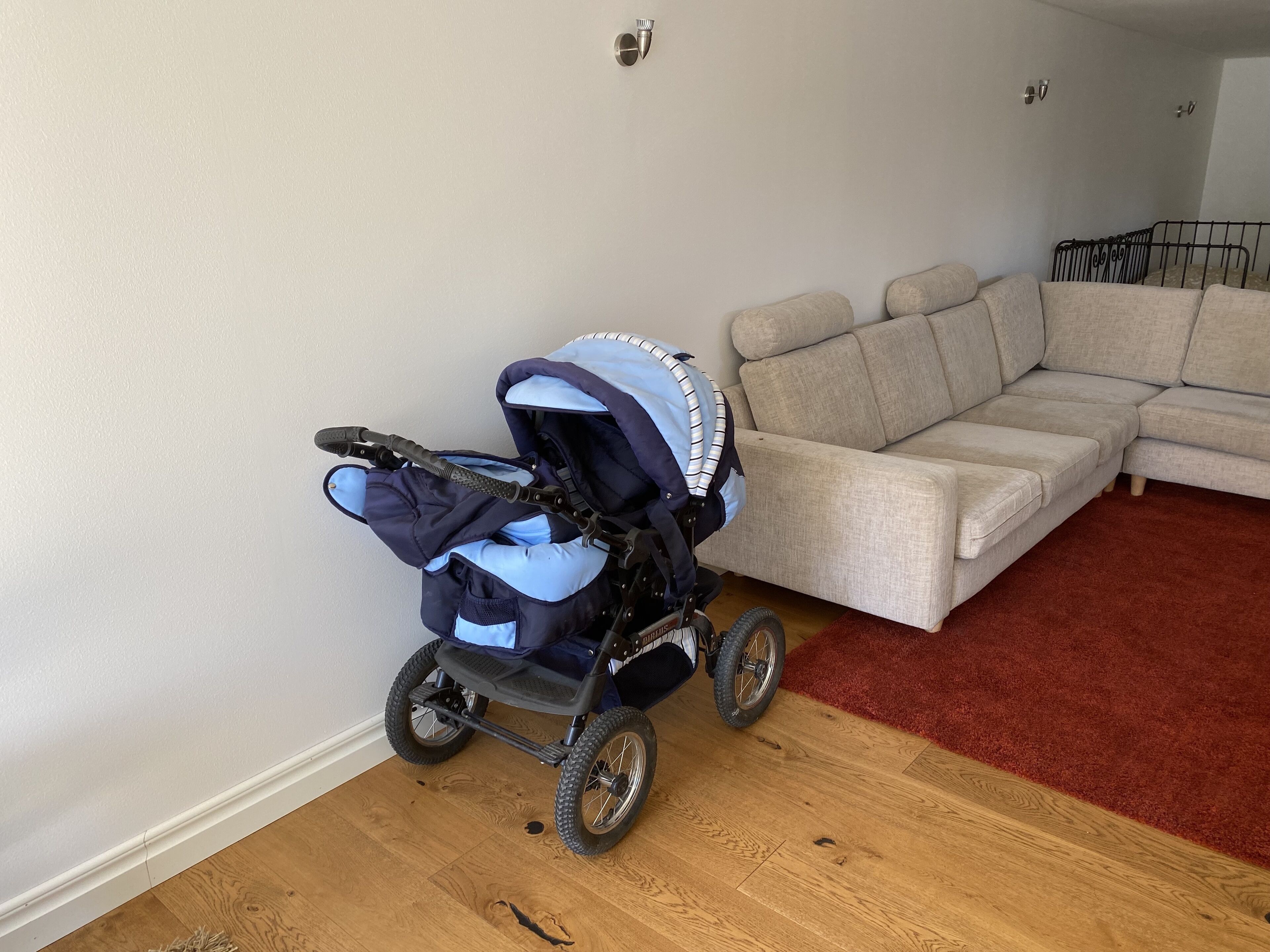 Une pièce avec un canapé d’angle beige, une moquette rouge et une poussette pour bébé.
