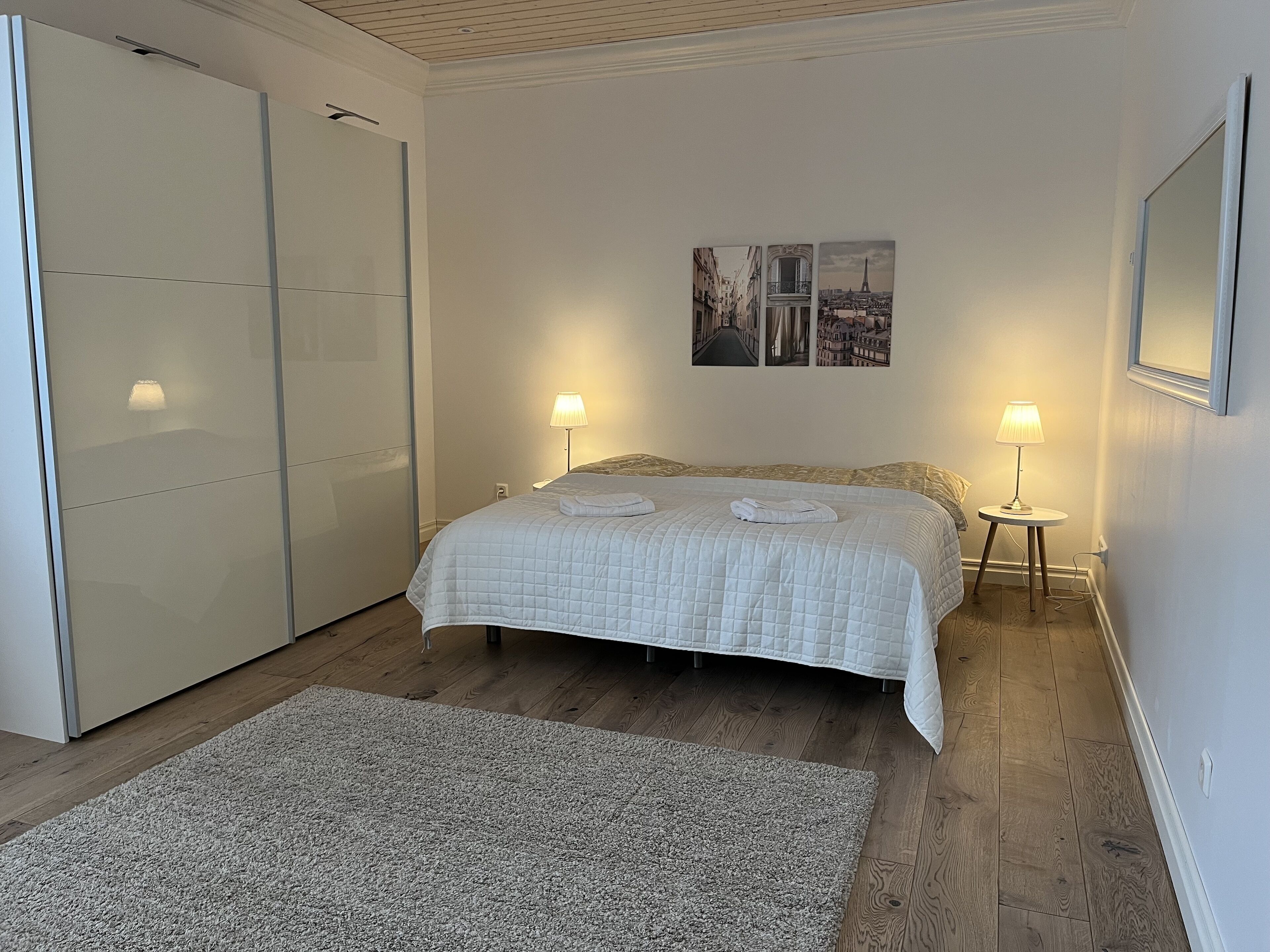 Een slaapkamer met een bed, twee bedlampen, een grote spiegel en een kunstwerk van een stadsgezicht aan de muur.