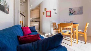 A living room with a blue sofa, red cushions, a dining table with chairs, and a kitchen in the background. - Apartment with swimming pool "Tivoli" (Bibione)