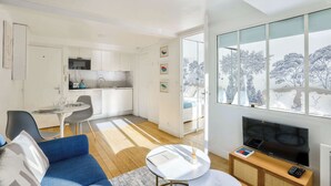 Een moderne woonkamer met een bank, een salontafel en een televisie. Achtergrond is een keuken met witte kasten en een eethoek met een tafel en stoelen.