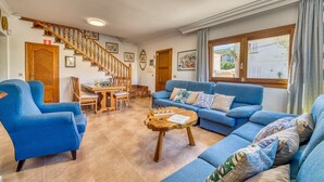 A spacious living room with a blue sofa set, a wooden dining table with chairs, a wooden staircase, and a large window with curtains. - House in Puerto Pollensa Villa Pintor with pool (Puerto Pollensa)