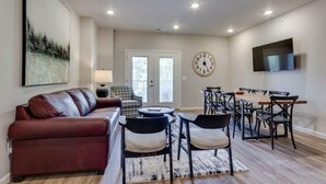 A living room with a red sofa, a dining area with a wooden table and chairs, a clock on the wall, and a TV mounted above the table. - SAVE! 2 Min to Pkwy! Luxury Apt Best Price (Sevierville)