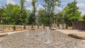 A fountain with water jets surrounded by a pebble-tiled area, a metal fence, and trees in the background. - SAVE! 2 Min to Pkwy! Luxury Apt Best Price (Sevierville)