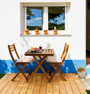 Une table et des chaises en bois sont disposées sur une terrasse en bois, avec une fenêtre au-dessus et une plante en pot sur le côté.