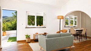 Ein Wohnzimmer mit einer Couch, einem Esstisch, Stühlen, einer Lampe und Blick auf Grün hinter den Fenstern.