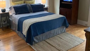 Un dormitorio con una cama, dos mesitas de noche, un televisor sobre un mueble bajo y una ventana con persianas.