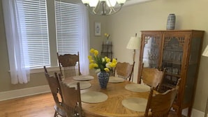 Comedor con una mesa redonda de madera, seis sillas, un florero con flores amarillas, una araña y un aparador con puertas de vidrio.