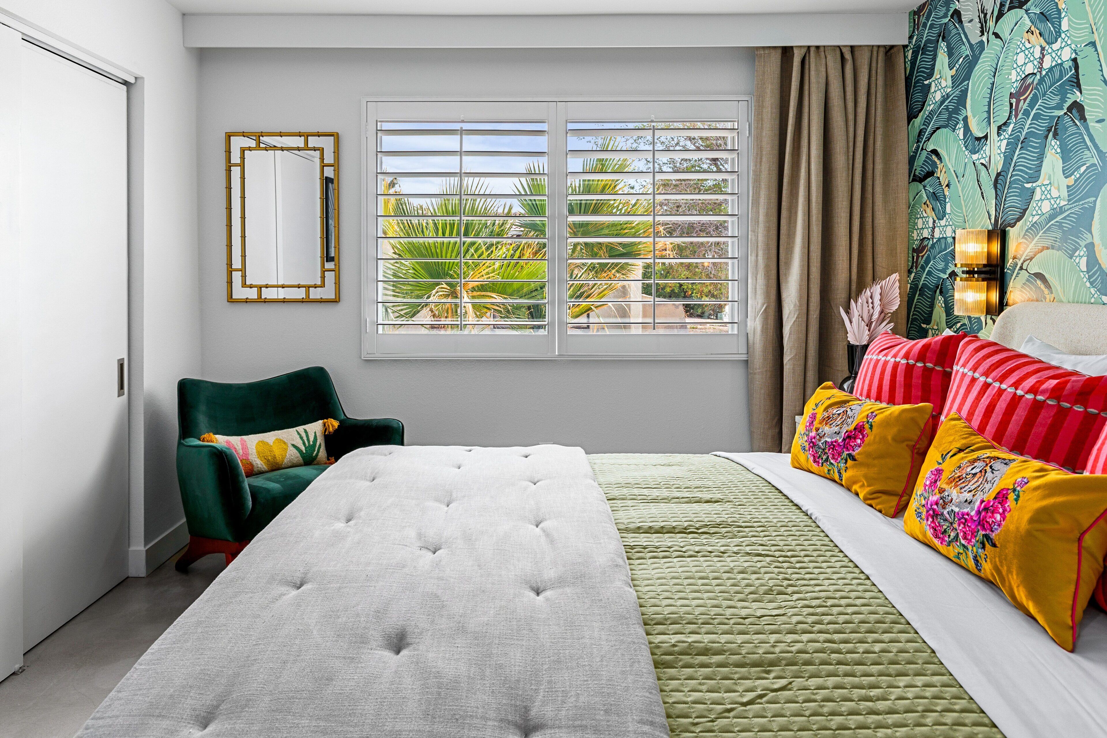 Une chambre avec un lit, une chaise, une fenêtre avec des stores et un mur orné d’un motif tropical.