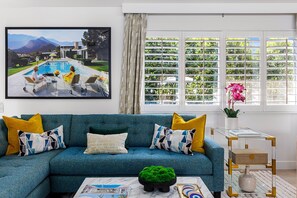 Una sala de estar con un sofá azul, una mesa de centro con revistas, un cuadro de una piscina y una ventana con vistas a follaje.
