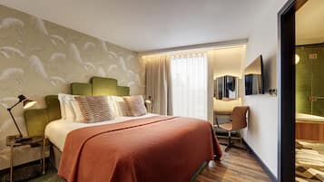 Habitación de hotel con cama, escritorio, silla, espejo y baño con ducha e inodoro.