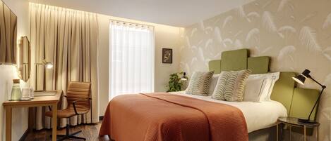A hotel room with a bed, a desk, a chair, a lamp, and a plant.