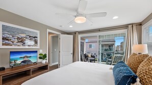 A hotel room with a bed, a TV on a wooden stand, a ceiling fan, a balcony with a table and chairs, and a view of buildings. - Pacific Surf  in Mission Beach | Steps to the Sand (San Diego)