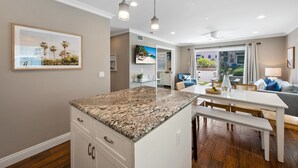 A modern kitchen with granite countertops, a dining area with a white table and bench, and a living area with a sofa and a TV. - Pacific Surf  in Mission Beach | Steps to the Sand (San Diego)