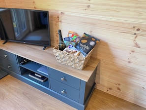 Habitación moderna con pared de madera, un televisor sobre un mueble de madera, un armario gris con cajones y una cesta de mimbre con aperitivos y una botella.