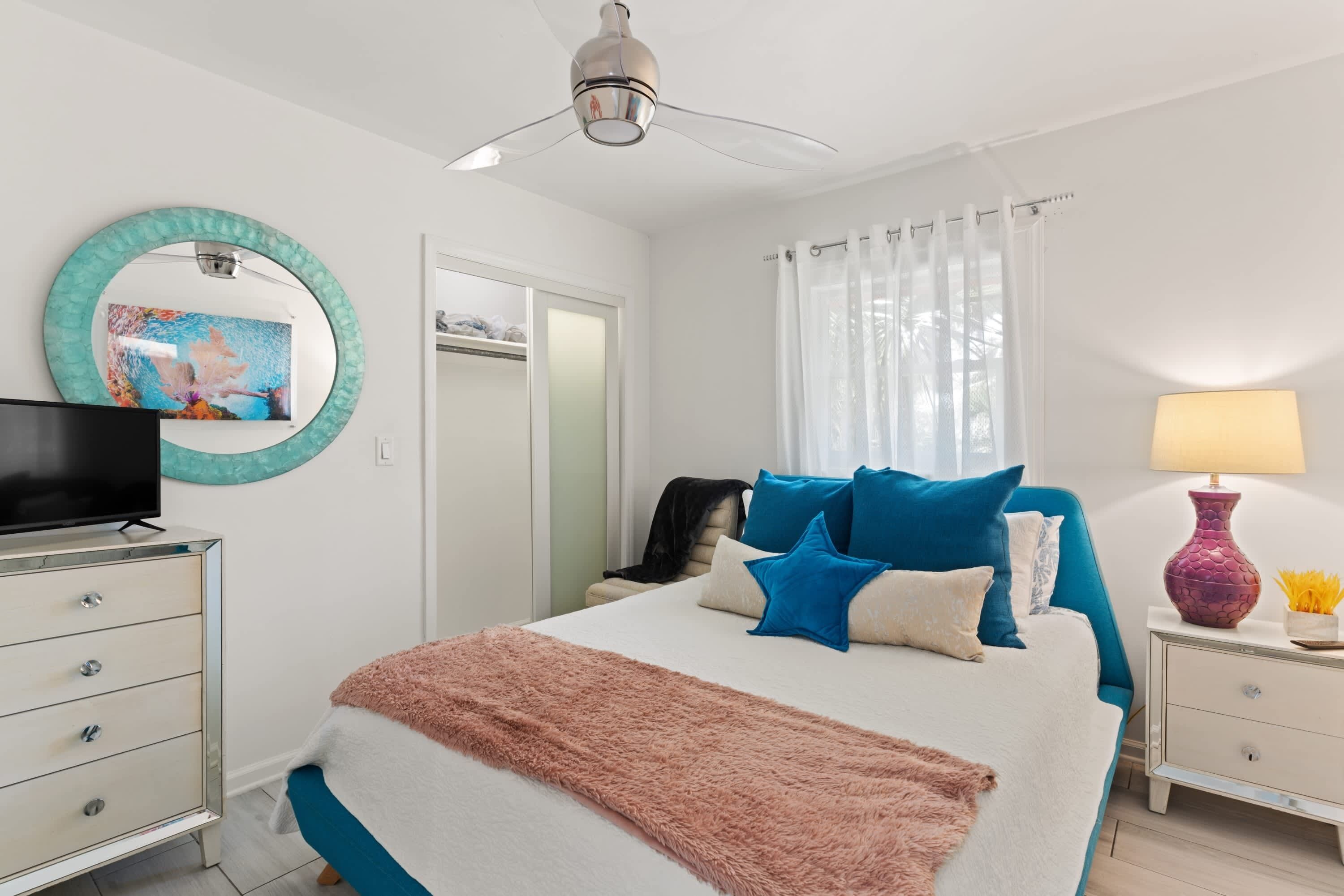 Une chambre à coucher comprenant un lit, un téléviseur, un ventilateur de plafond, un miroir, une armoire et une table de chevet avec une lampe.