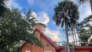 Una casa a due piani con tetto rosso e un camino in mattoni, circondata da alberi e un cielo sereno.