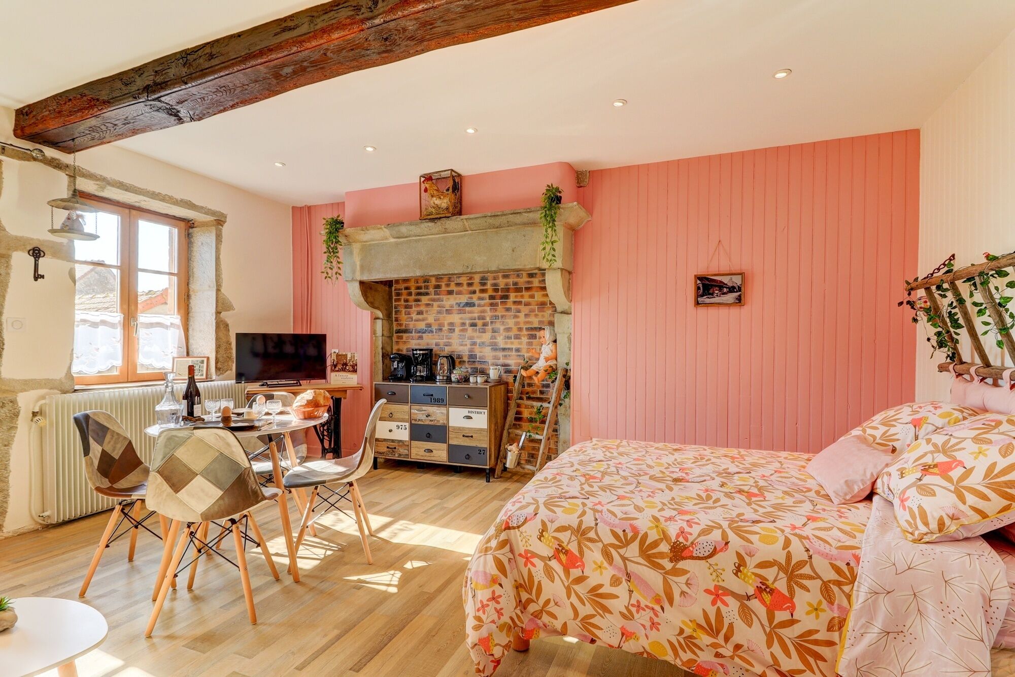 Una habitación acogedora con chimenea de ladrillo, un comedor con mesa y sillas, una cama con ropa de cama floral y una pared rosa.