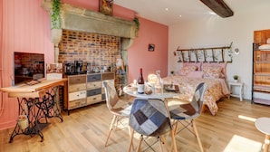 A cozy room with a brick fireplace, a bed with floral bedding, a dining table with chairs, and a vintage-style desk with a television. - Le Petit Pressoir - Charmant studio (Collonge-en-Charollais)