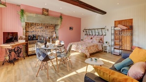 A cozy room with a brick fireplace, a bed with floral bedding, a dining area with a table and chairs, and a wooden ceiling. - Le Petit Pressoir - Charmant studio (Collonge-en-Charollais)