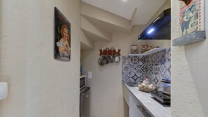 A narrow kitchen with a microwave, stove, and hanging pots. A decorative plate hangs on the wall, and a patterned tile backsplash is visible. - Le Petit Pressoir - Charmant studio (Collonge-en-Charollais)